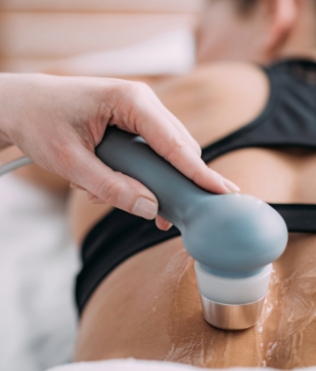 Close-up of a hand holding a skincare device applied to a person's back, who is wearing a black top and lying face down, with gel or oil for smooth movement, focusing on relaxation and body care against a softly blurred background.
