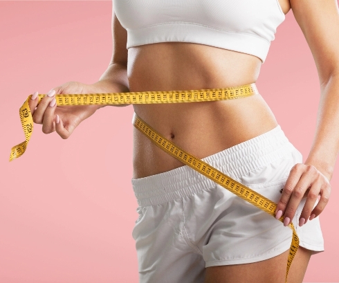 Close-up of a woman measuring her waist with a yellow tape measure, dressed in white sports attire against a soft pink background, emphasizing health, fitness, and self-improvement