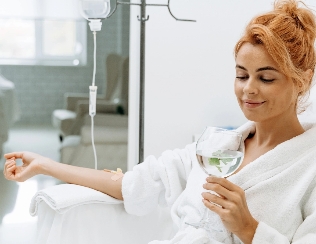 Woman in a white bathrobe sitting comfortably with a relaxed expression, holding a glass of water with lemon or lime, receiving an IV drip treatment, in a tranquil and minimalistic setting, emphasizing wellness, self-care, and relaxation.